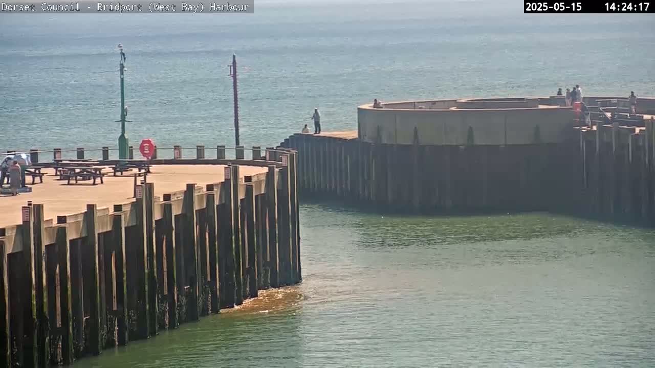 A harbor with a concrete pier and wooden pilings, a few people are visible, under hazy sunny conditions.