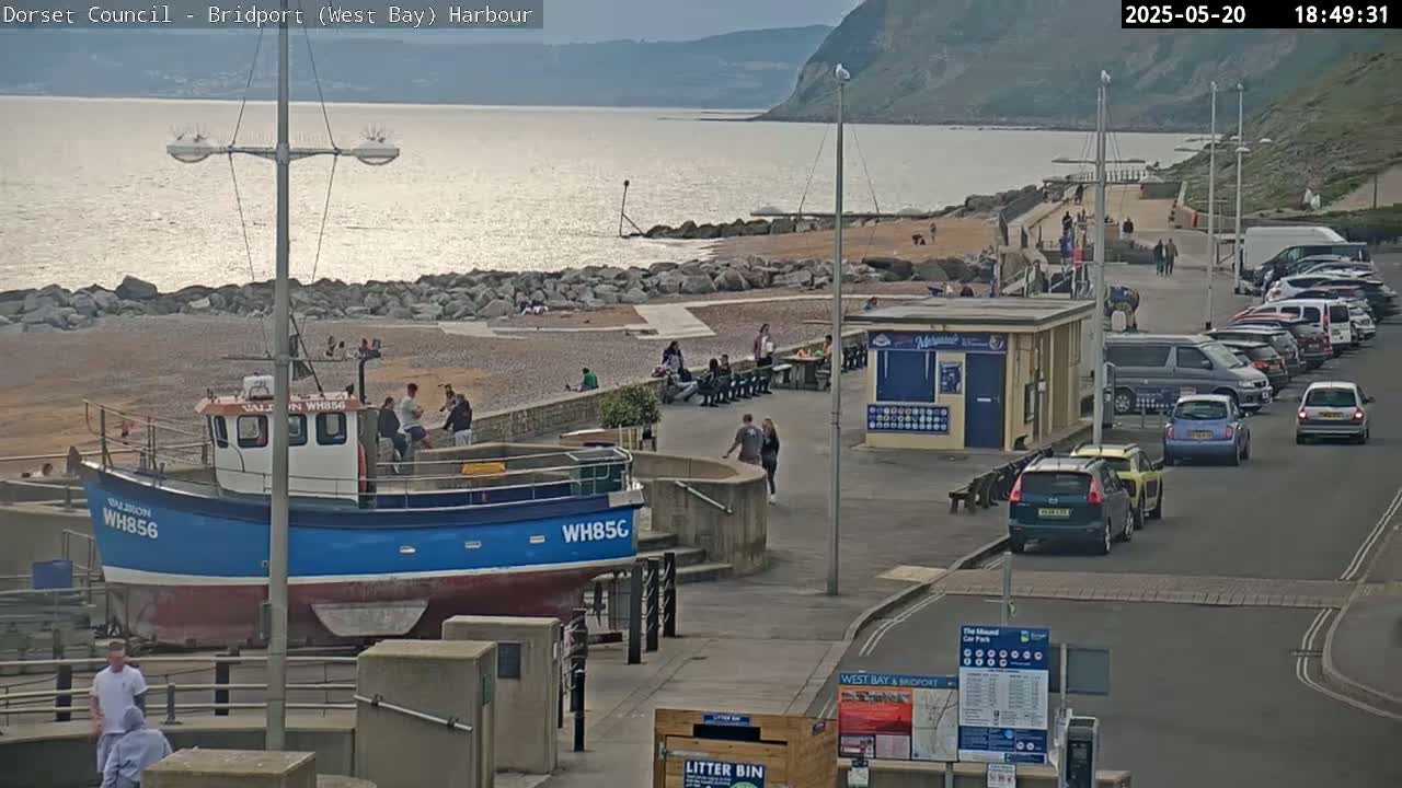 A blue and white fishing boat is docked near a pebble beach with people relaxing and walking, a calm sea, and cars parked in a lot next to a building, all under a partly cloudy sky.