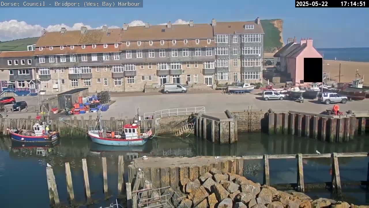 A harbor scene on a sunny day shows several small fishing boats moored in calm water near a multi-story building and a sandy beach.