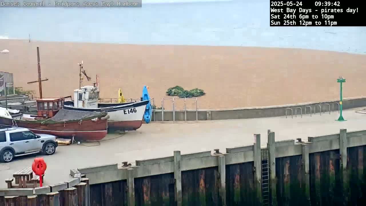 A harbor scene shows several small boats docked near a sandy beach under an overcast sky.