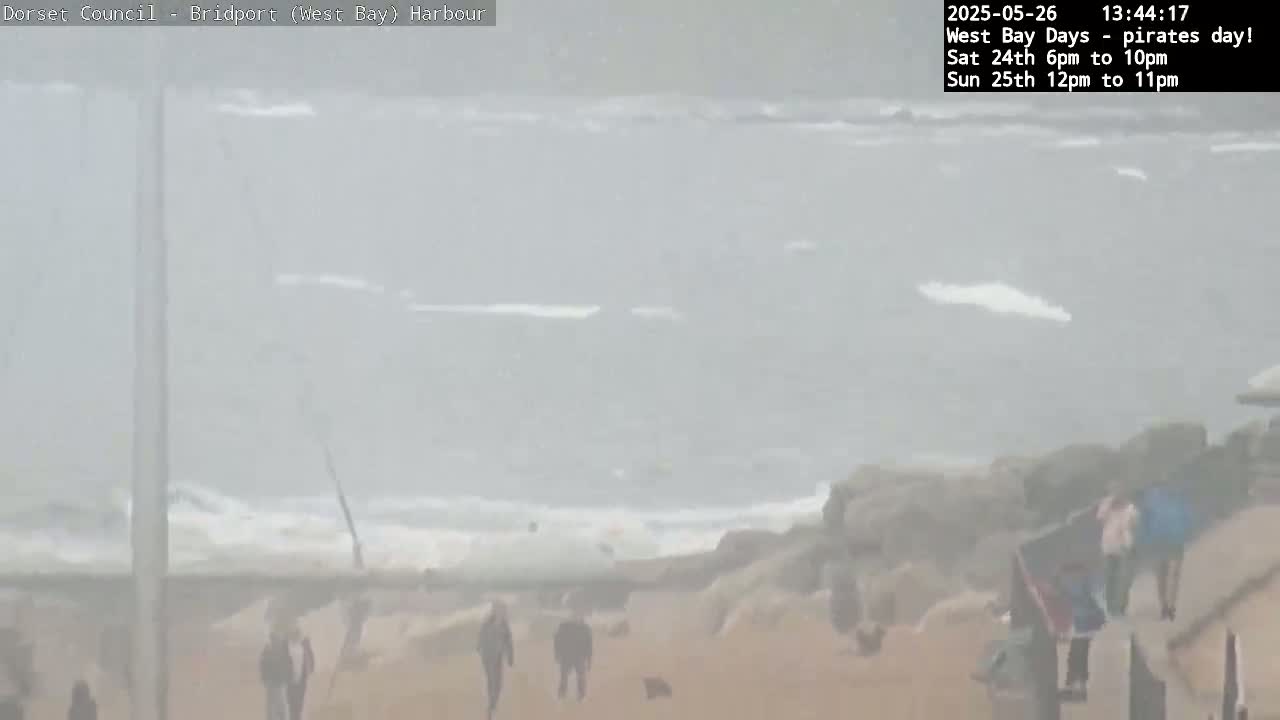 A hazy, overcast day at a beach shows a gray sea with small waves crashing on a rocky shore where several people are walking along the sand.