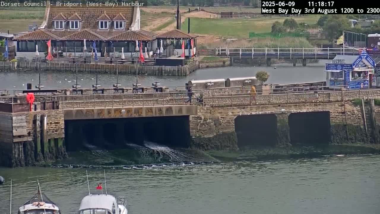 Water flows under a stone bridge near a harbor, with a restaurant and small building visible in the background under a cloudy sky.