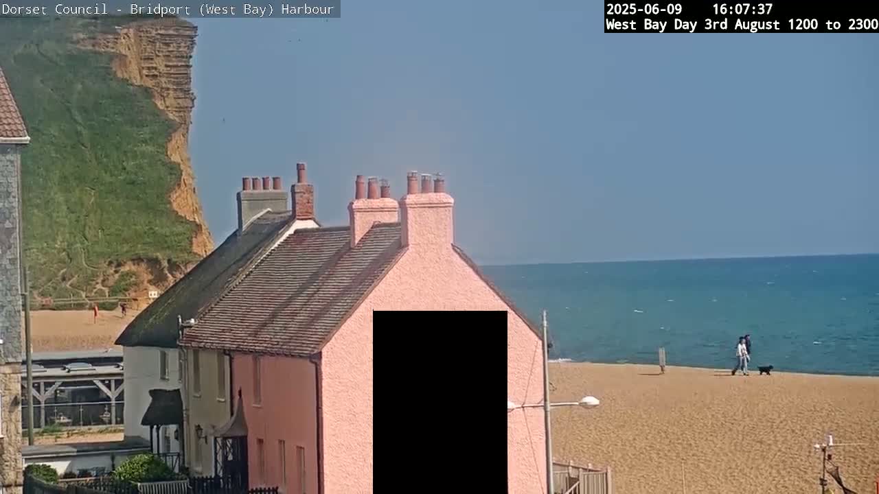 A pink house with a dark section obscuring a portion of its facade sits overlooking a sandy beach and calm ocean under a clear blue sky.