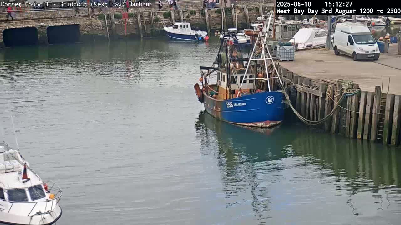 A blue fishing boat is docked at a harbor next to a smaller white boat, a van, and other boats, on a calm day.