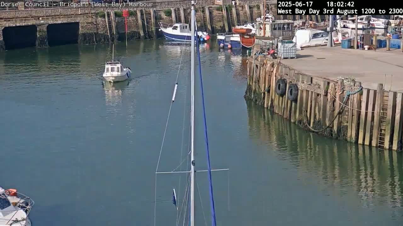 A sailboat mast is centered in a harbor with several smaller boats moored nearby on a sunny day.