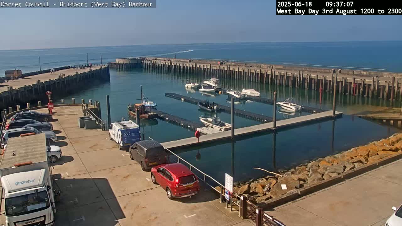A harbor with several boats docked, cars parked in a lot, and calm blue water under a sunny sky.