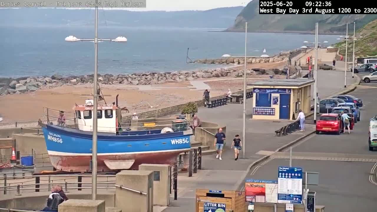 A blue fishing boat sits on land next to a paved walkway overlooking a calm ocean beach on a sunny day.