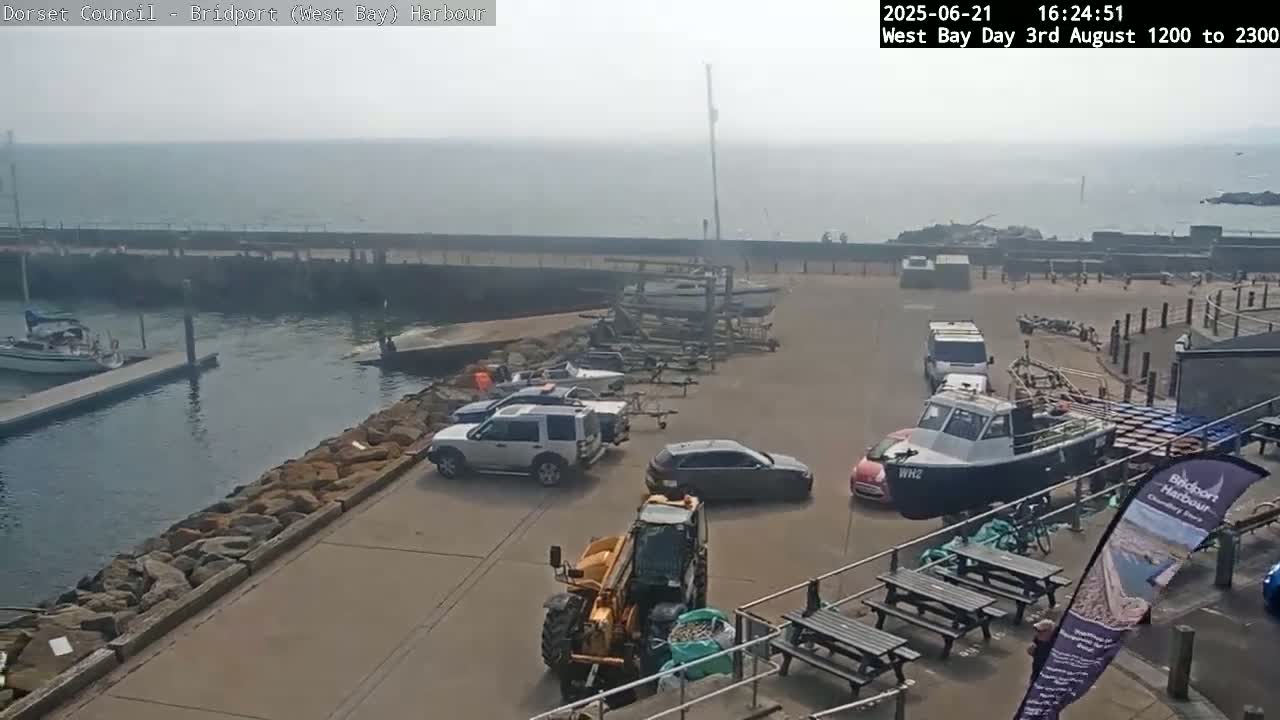 A harbor scene on a hazy day shows several cars and a small tractor parked near a boat on a trailer; a sailboat is docked in the water beyond a stone seawall.