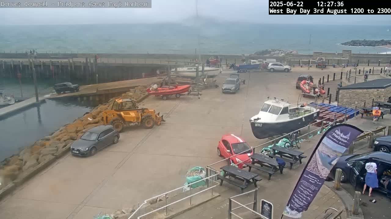 A harbor scene on an overcast day shows several cars, boats, and a small construction vehicle parked near a stone wall and waterfront.