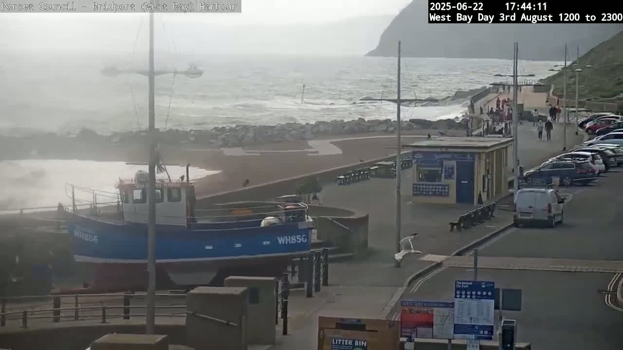 A small blue and white fishing boat is docked near a harbor walkway with people walking by, amidst a wavy, somewhat stormy sea.