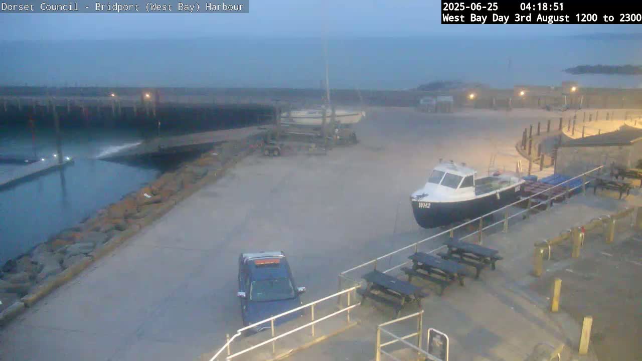 A harbor at dawn shows a small fishing boat and a vehicle parked near picnic tables, with calm water and a sailboat in the background.