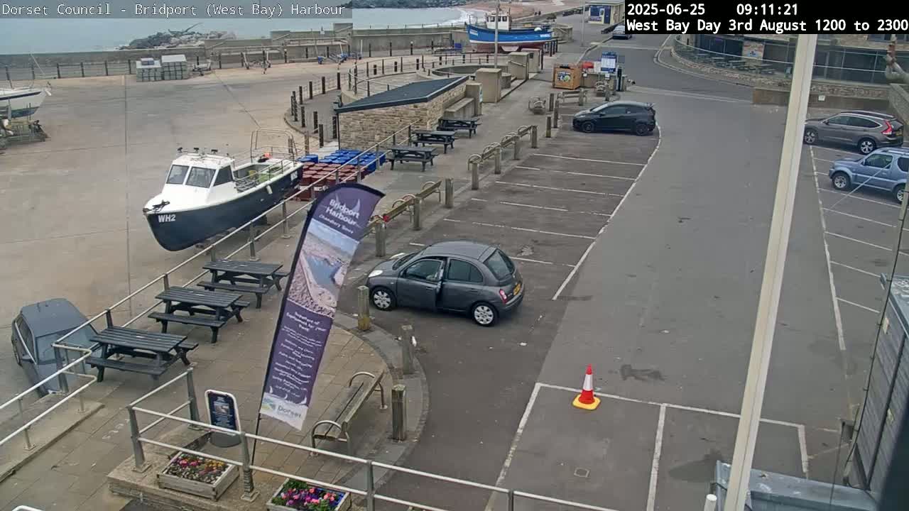 An overcast day at a harbor shows a parking lot with several cars, picnic tables, a small boat, and a partially visible larger boat in the background.