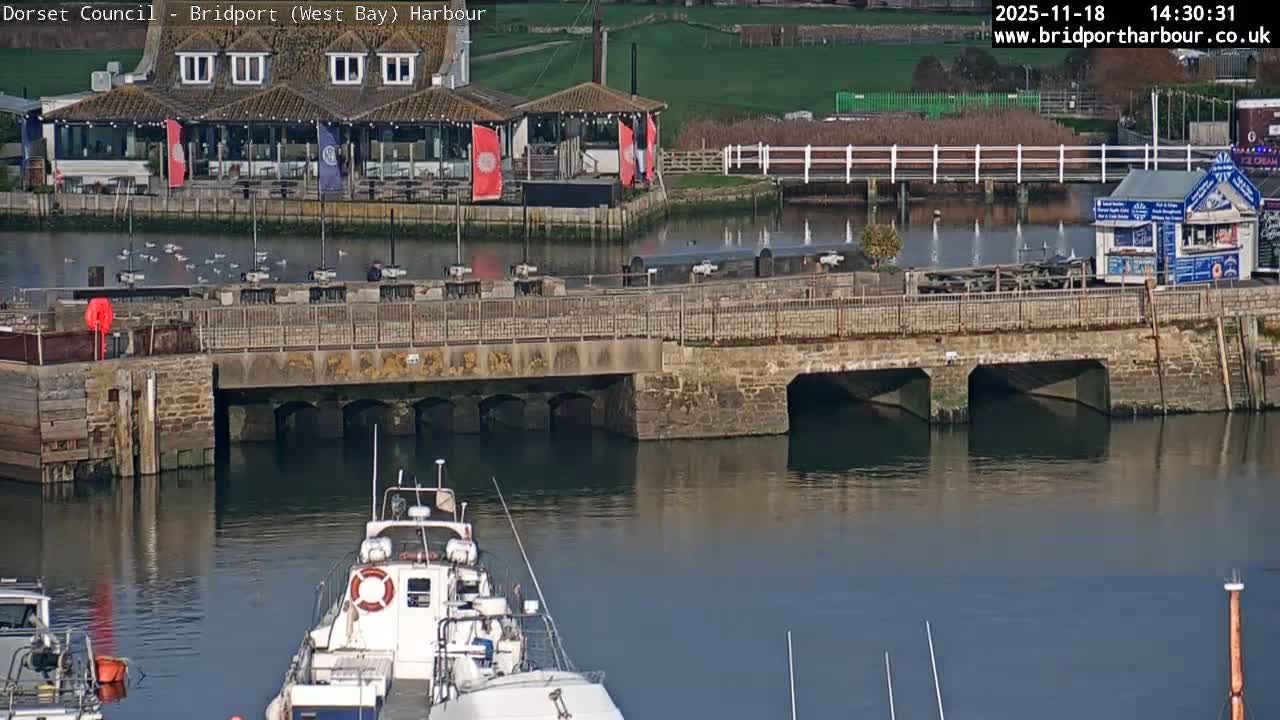 A harbor scene shows several boats moored near buildings, a sandy beach, and calm water under an overcast sky.