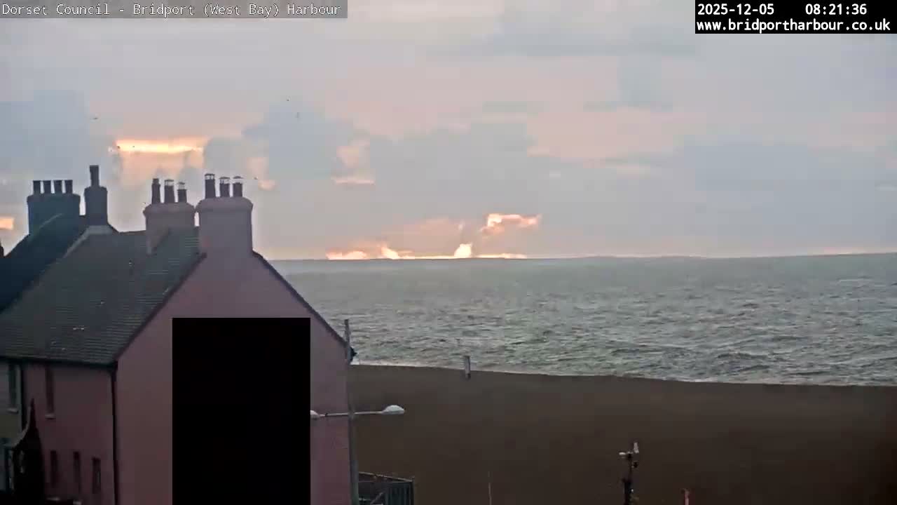 Coastal rooftops and chimneys frame a moderately choppy sea under an overcast sky, illuminated by a striking golden-orange glow from a cloudy sunrise on the distant horizon.