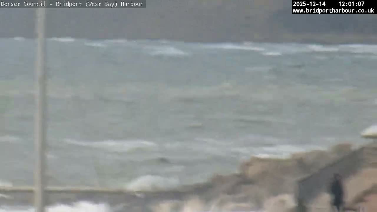 Coastal rooftops and chimneys frame a moderately choppy sea under an overcast sky, illuminated by a striking golden-orange glow from a cloudy sunrise on the distant horizon.
