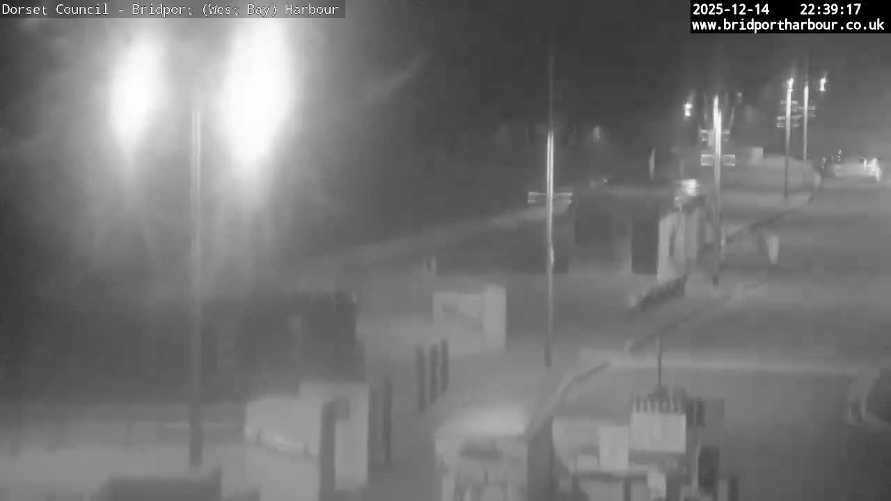 Coastal rooftops and chimneys frame a moderately choppy sea under an overcast sky, illuminated by a striking golden-orange glow from a cloudy sunrise on the distant horizon.