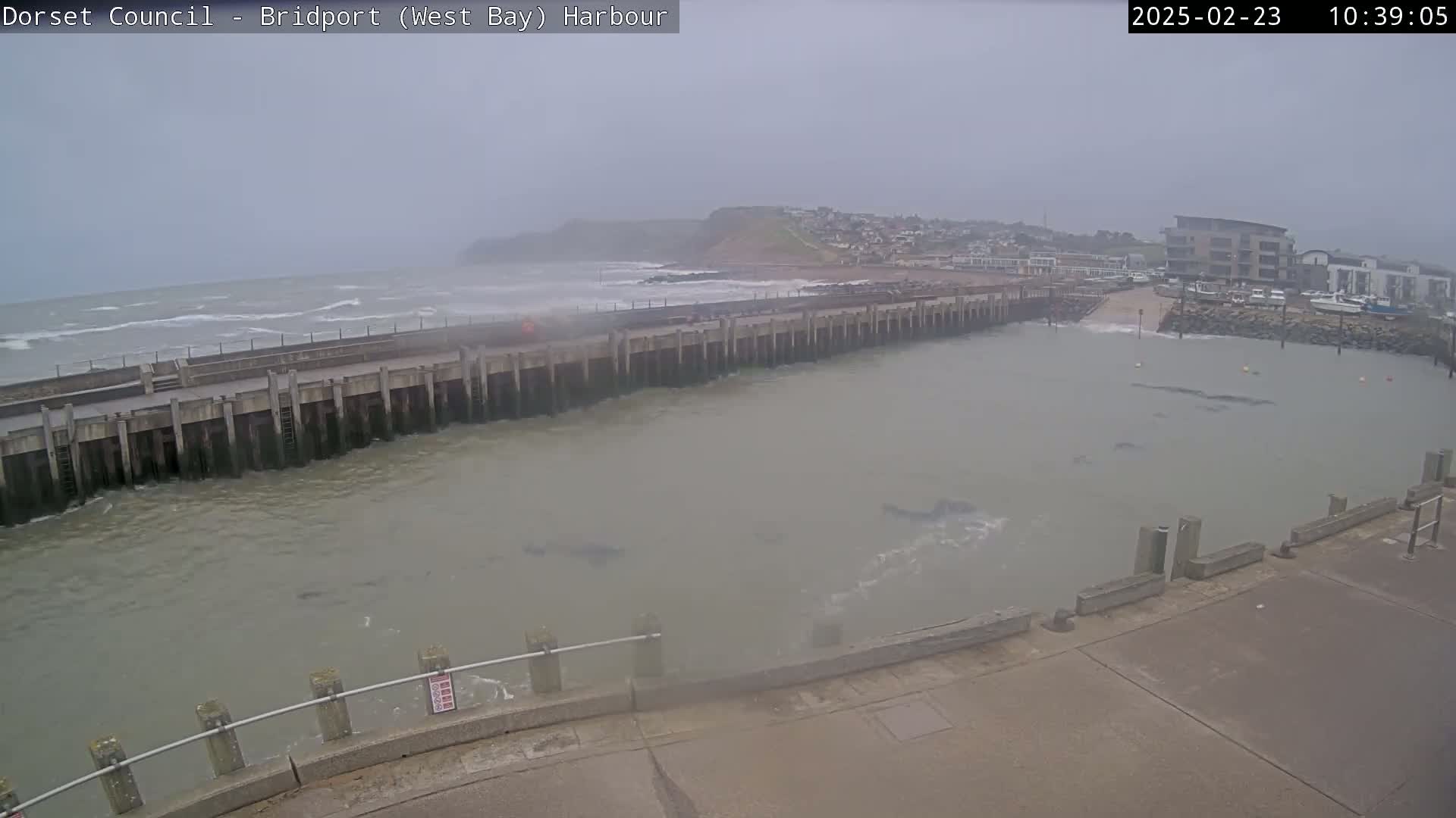 A harbor with a long concrete wall separating choppy ocean waves from calmer harbor waters under an overcast sky.