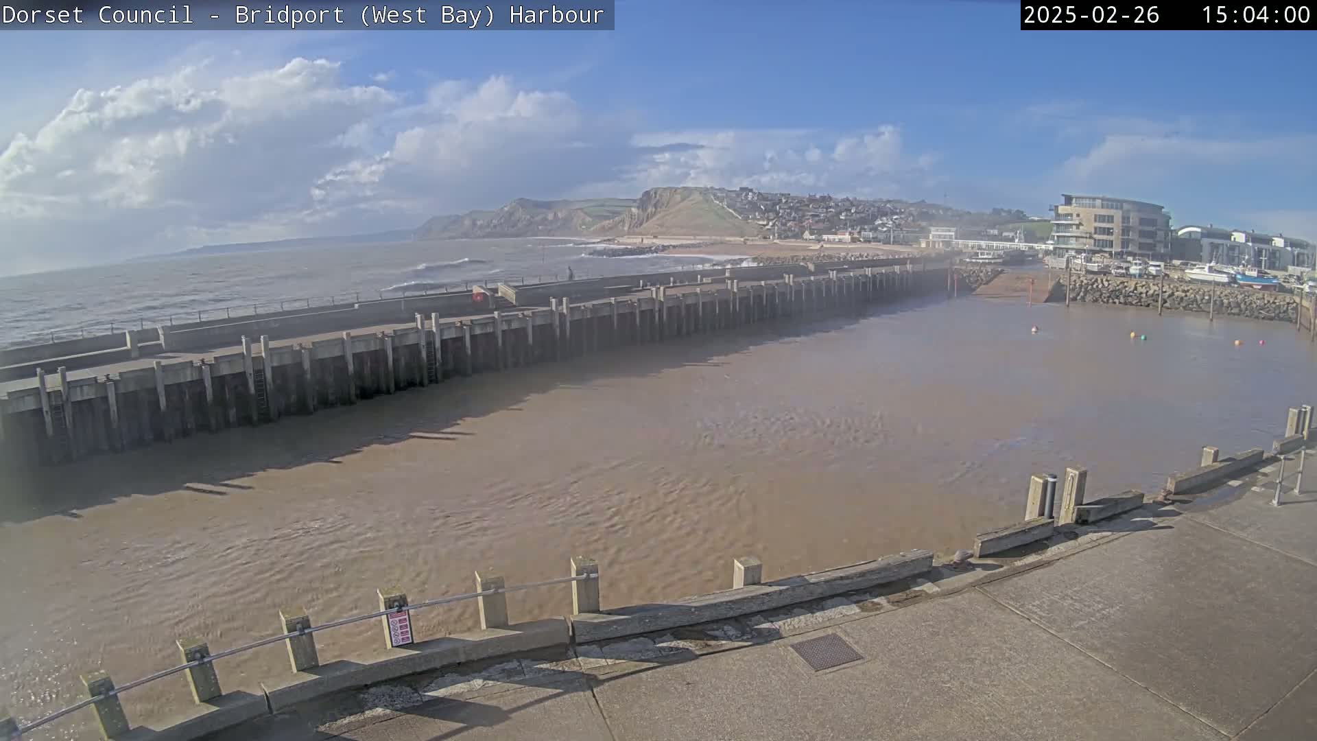 A harbor with calm, muddy water is partially enclosed by a long concrete wall, beyond which lies a wavy ocean under a partly cloudy sky.