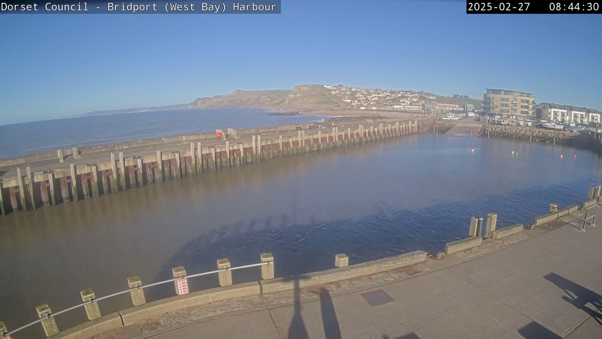 A harbor with calm, brown water is seen under a clear blue sky; a long, low wall separates the harbor from the ocean, with a town and cliffs visible in the distance.