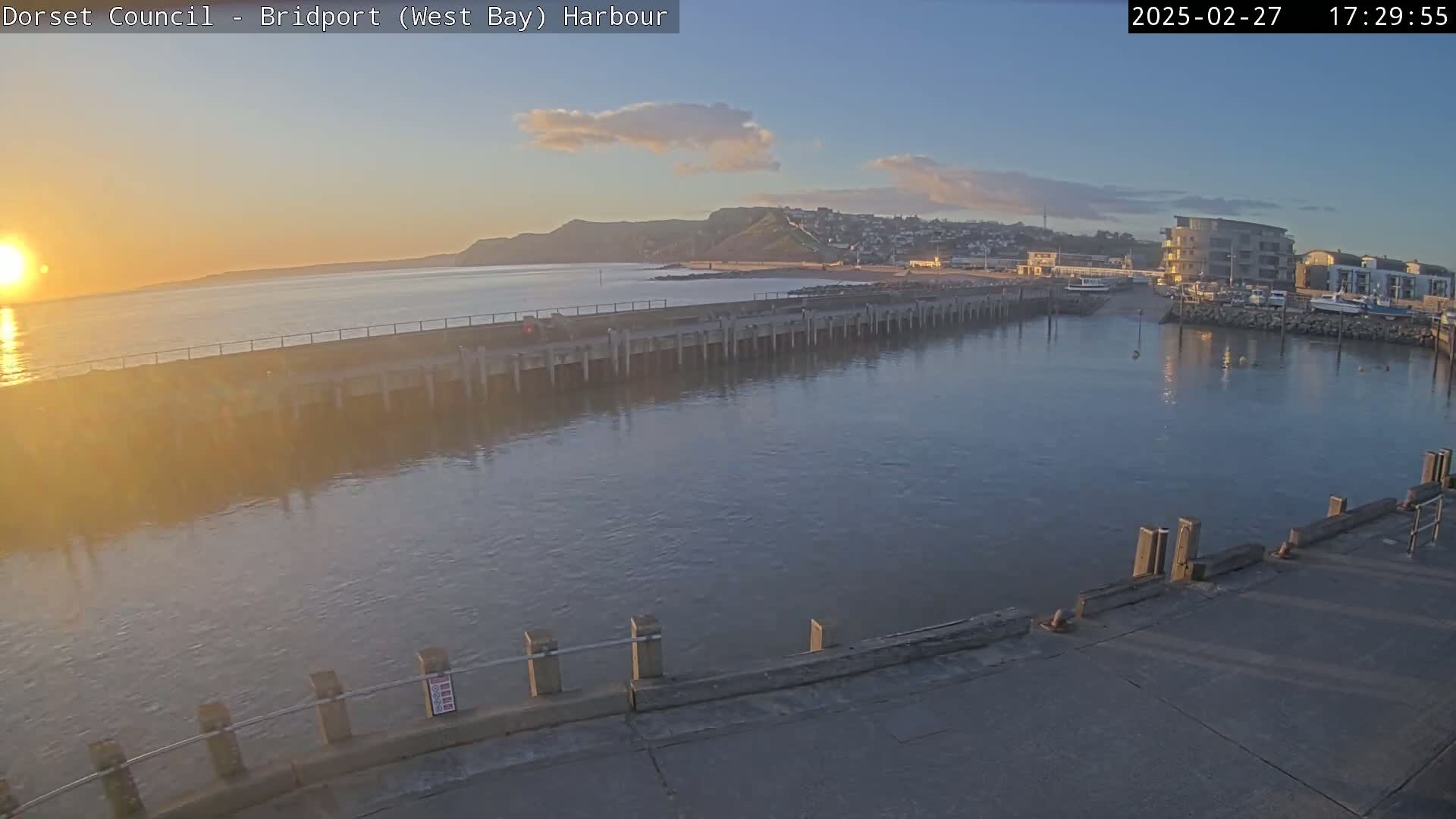 A harbor at sunset, with calm water reflecting the orange glow, shows a long pier extending into the bay near a hillside town.