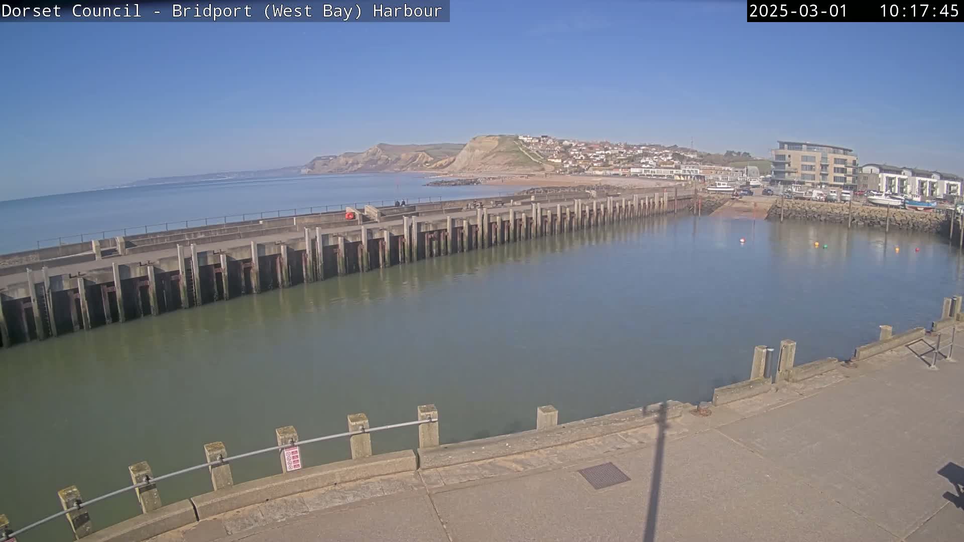 A harbor with a long concrete wall separating it from the sea, under sunny skies, shows a town and cliffs in the background.