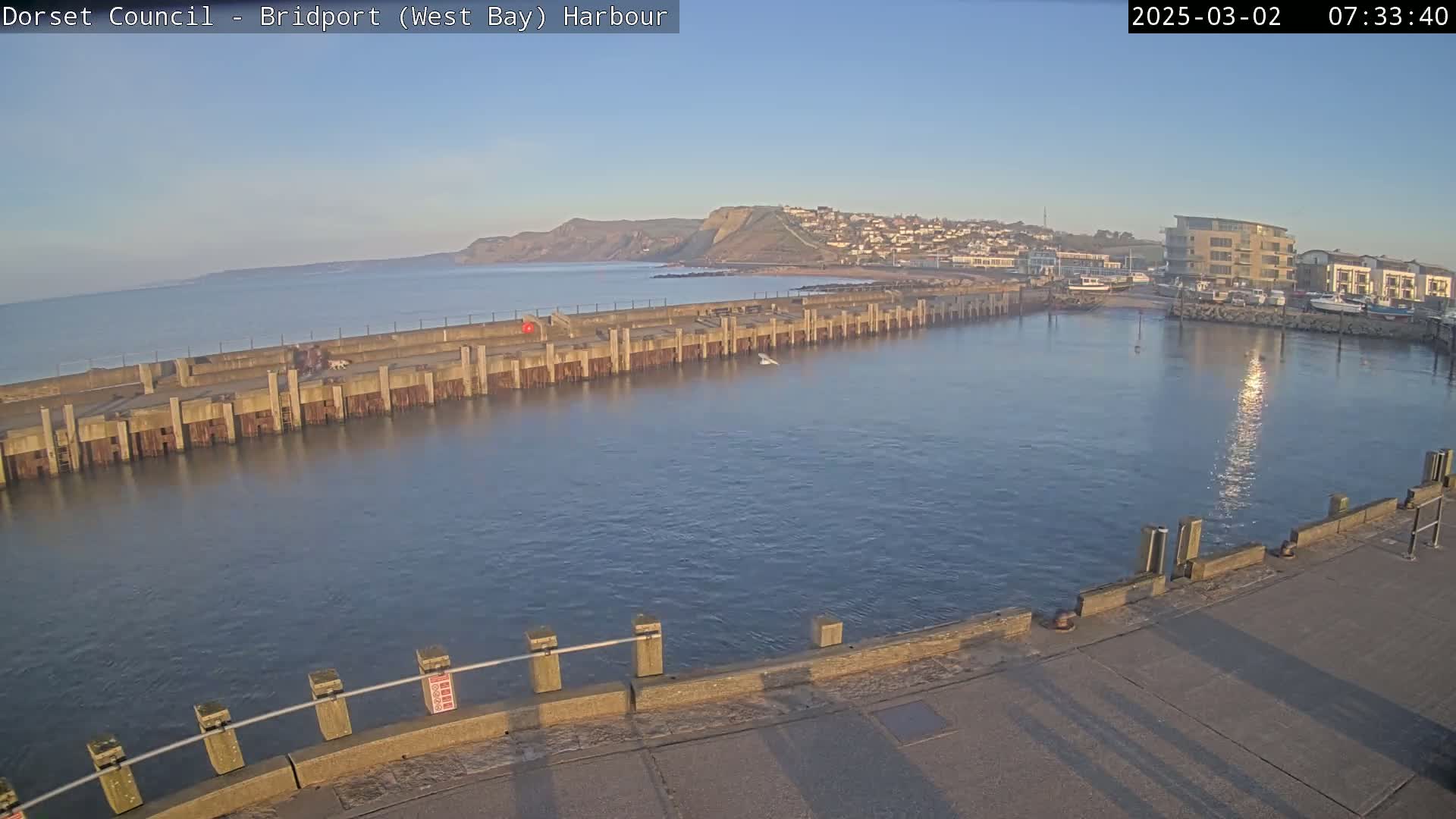 A calm harbor with a long concrete pier, a small town on a hillside in the background, and a clear, sunny sky.
