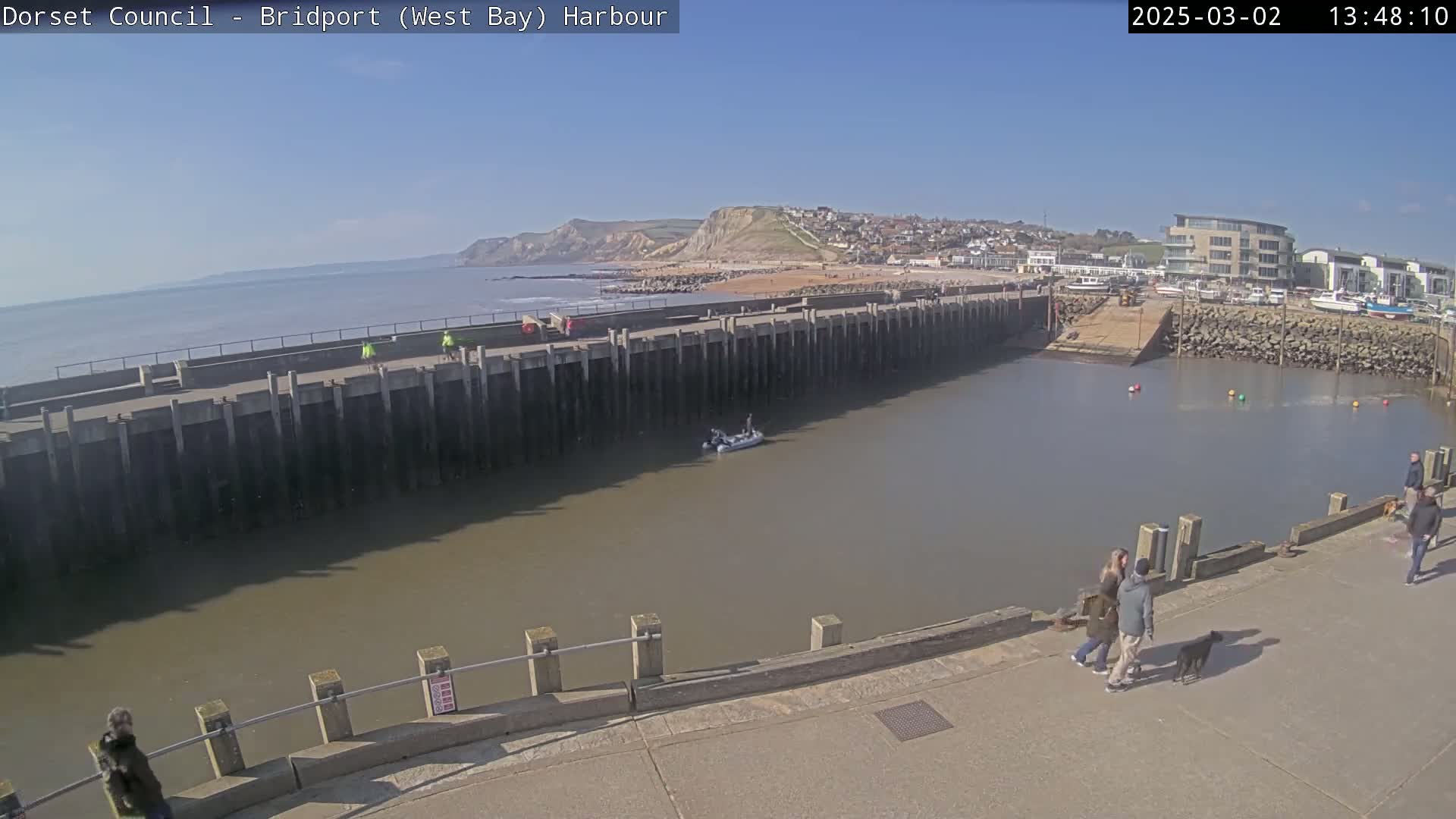 A harbor with calm, brown water is viewed from above on a sunny day, showing a small boat, people walking, and a coastal town with cliffs in the background.