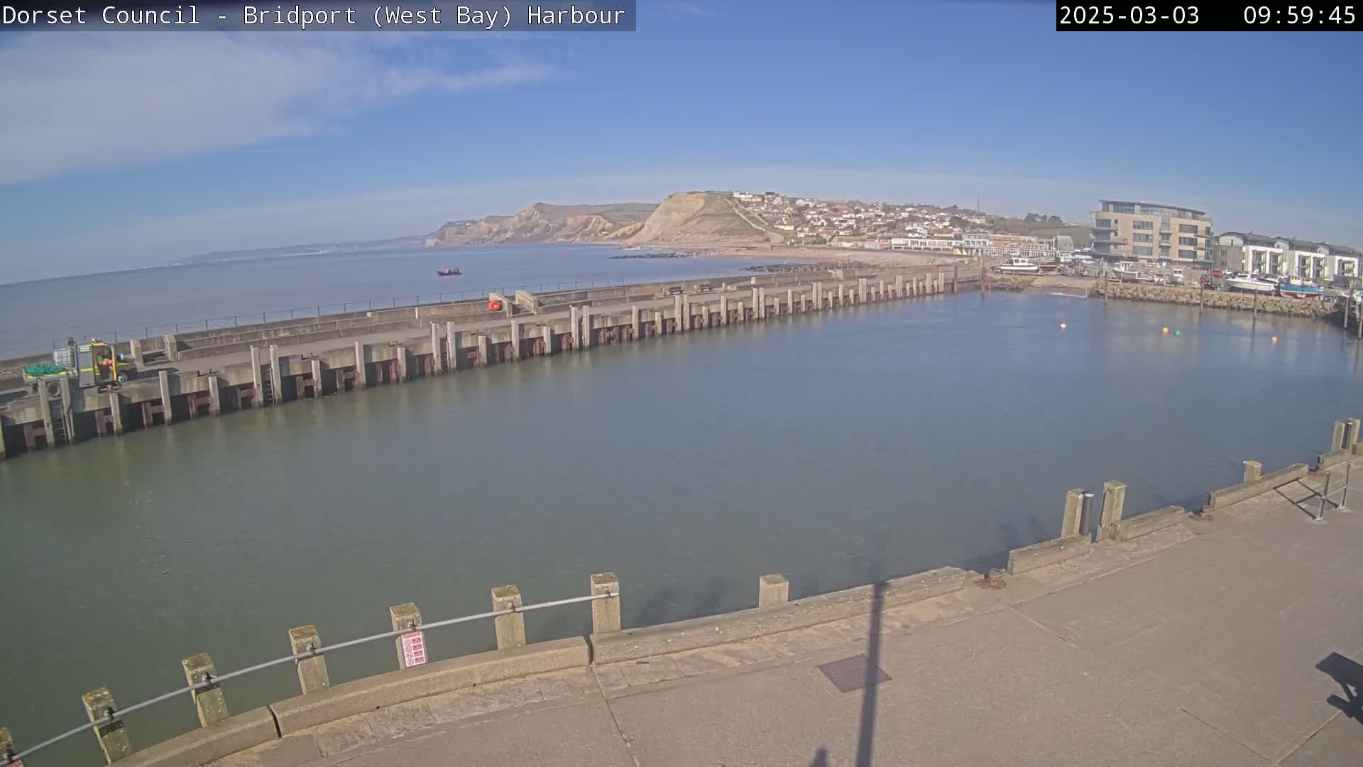 A harbor with calm water, a long concrete pier, and a small town nestled against a cliffside under a sunny sky.