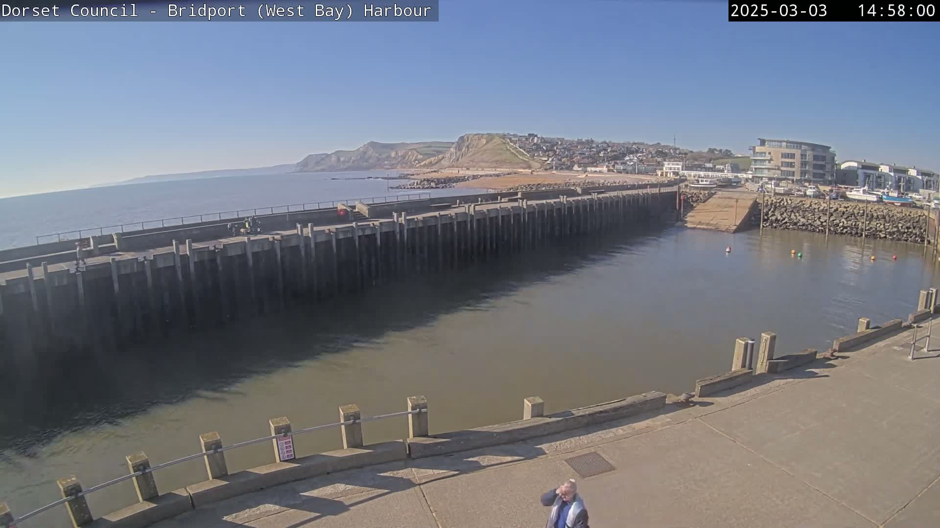 A harbor with a calm, muddy-brown sea under a clear blue sky shows a long concrete pier, a small town, and distant cliffs.