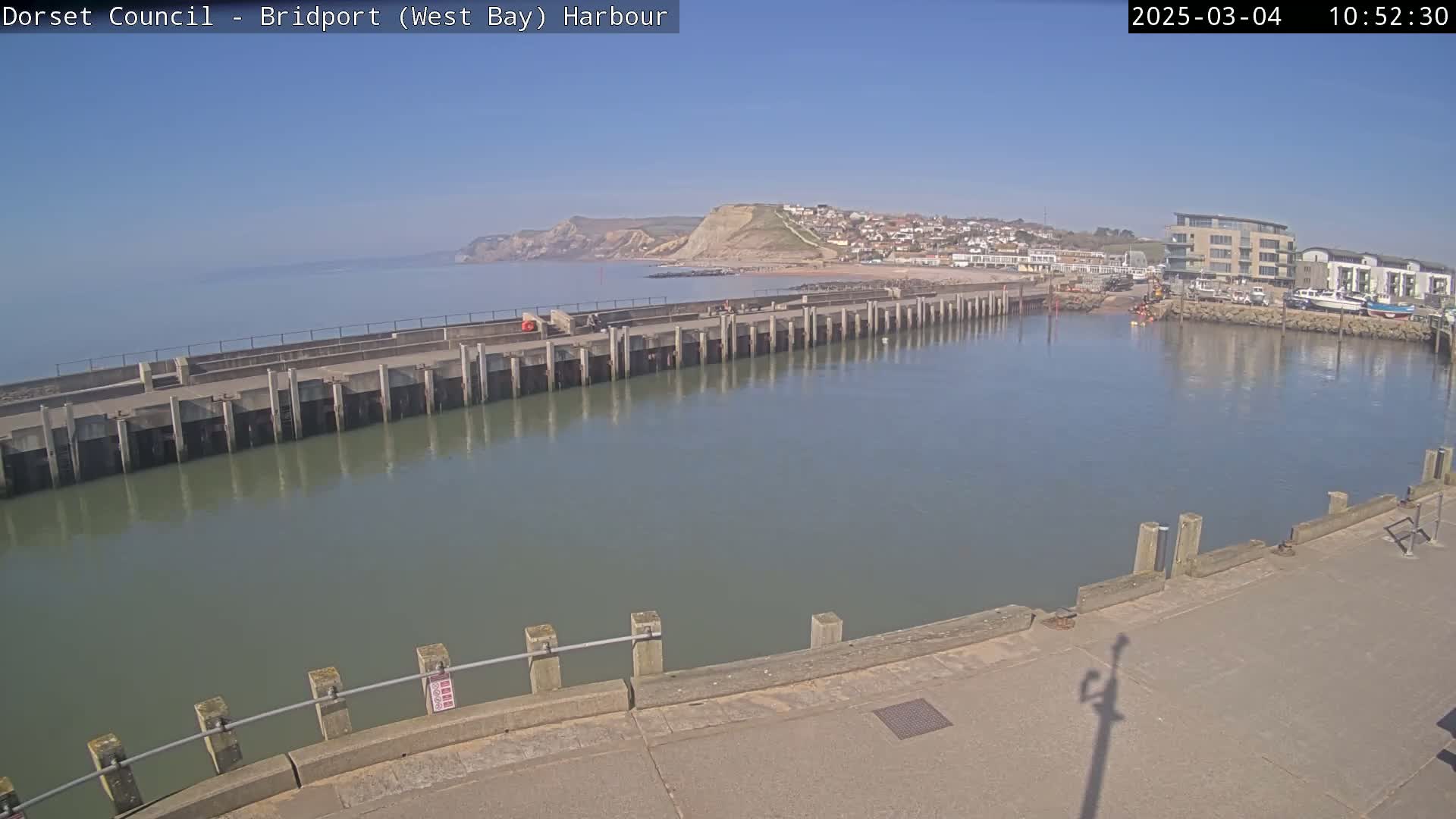 A harbor with a long concrete pier extends into calm, greenish-grey water under a clear blue sky, with a hillside town visible in the distance.