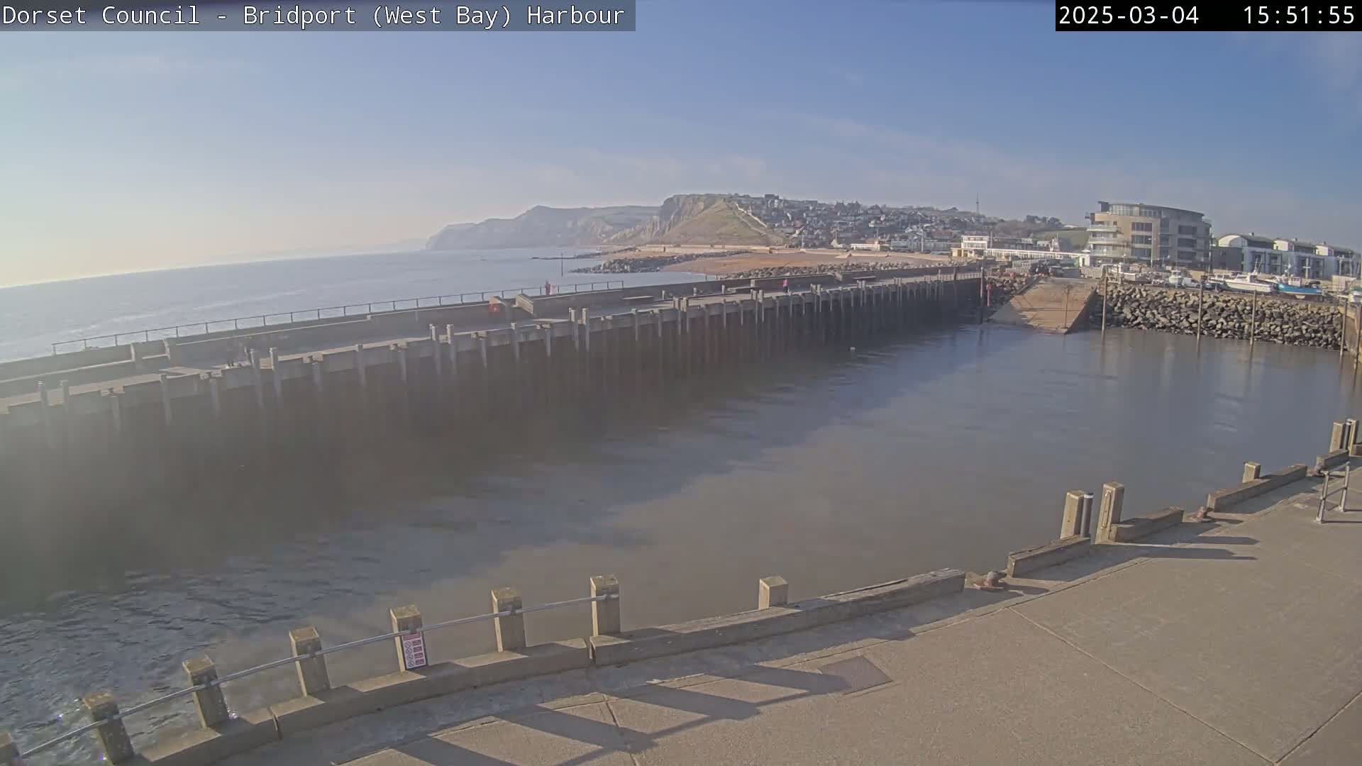A harbor with a long concrete pier extends into calm, gray water under a clear blue sky, with a sandy beach and cliffs visible in the distance.