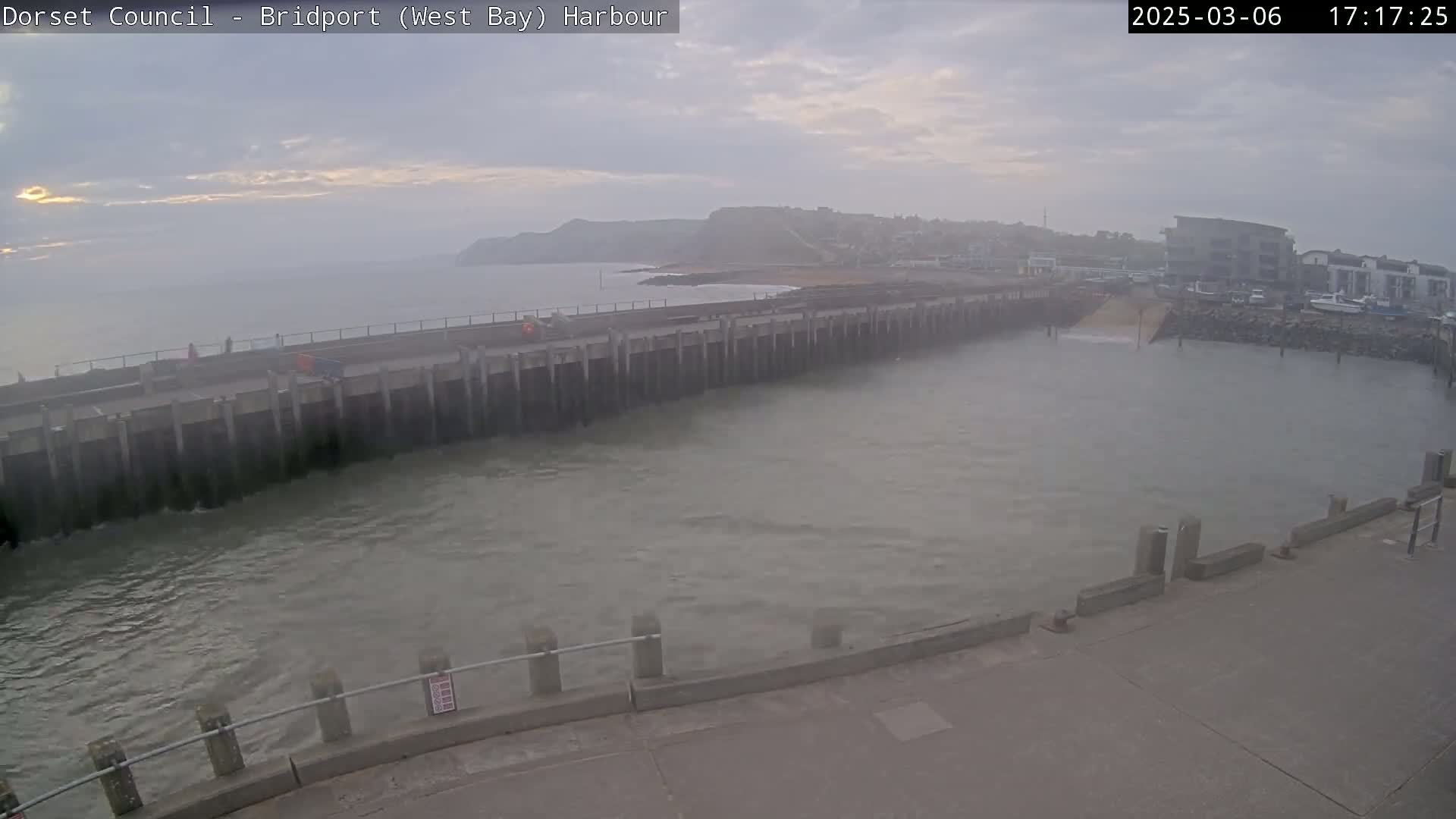 A hazy harbor scene shows a long concrete pier extending into a calm, grey body of water under an overcast sky.