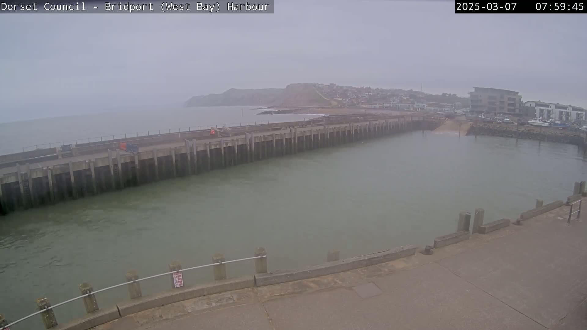 A hazy view of a harbor with a long concrete pier extending into calm, murky water, backed by a coastal town and cliffs.