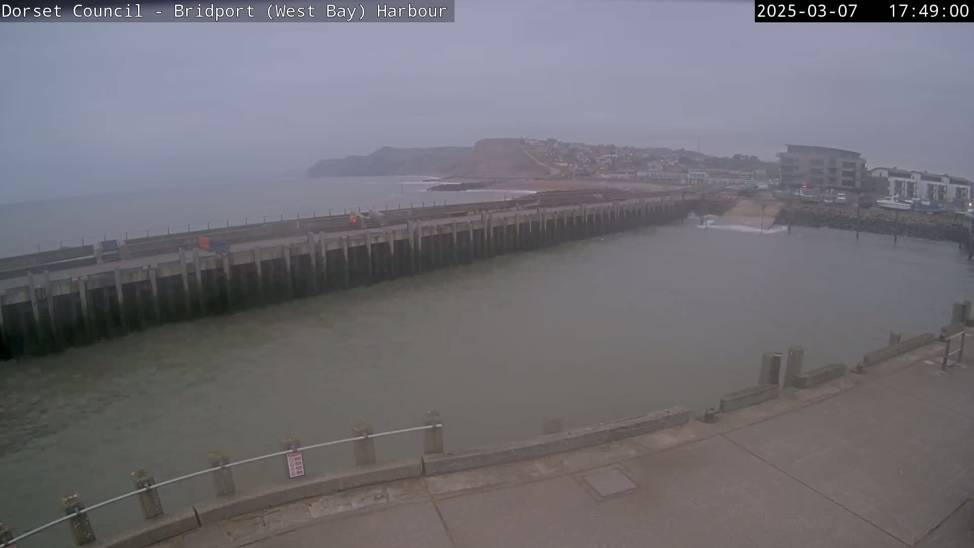 A harbor with a long concrete pier extends into calm, gray water under an overcast sky, with a small town and a cliff visible in the background.