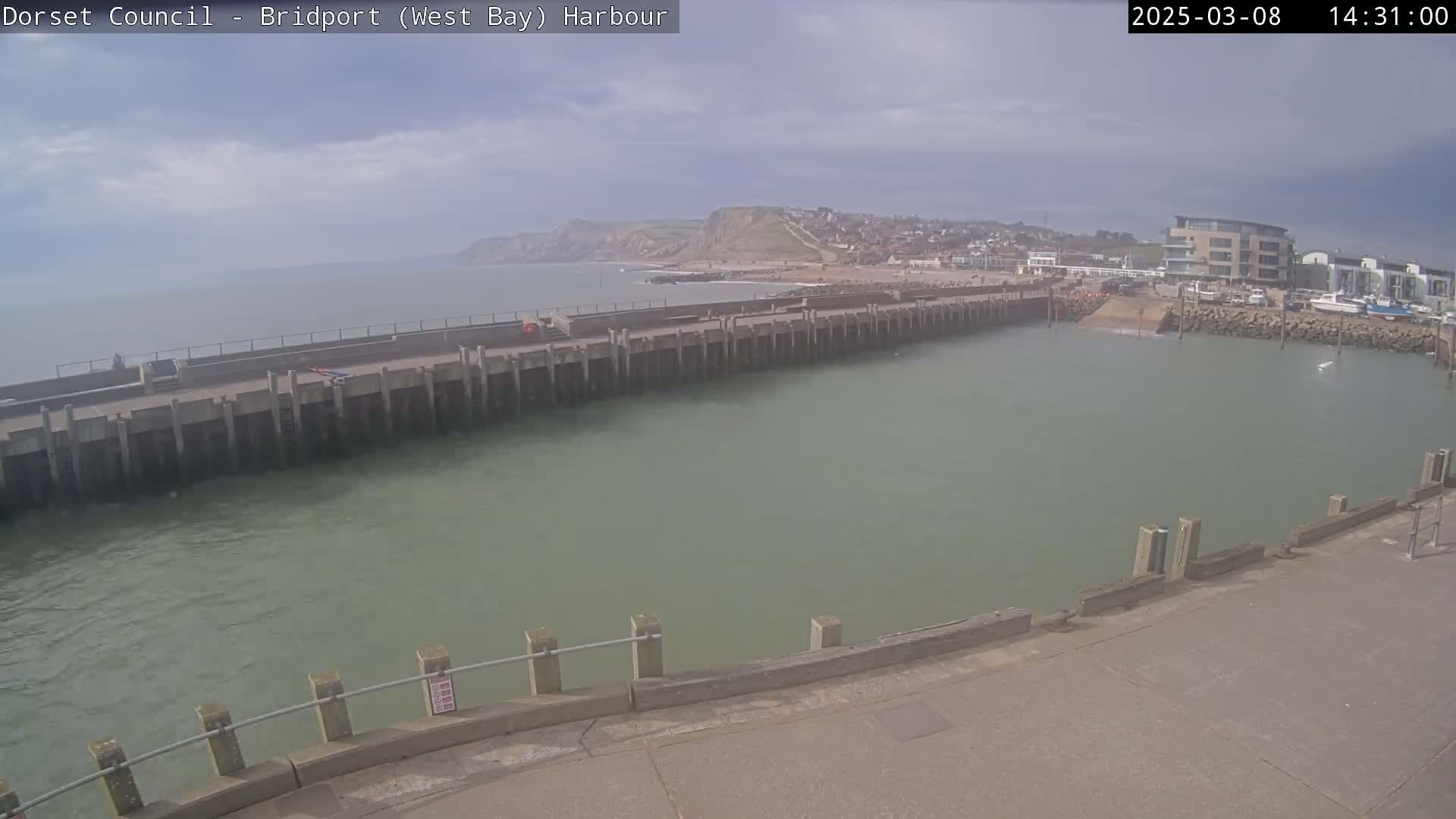 A harbor with calm, greenish water is partially enclosed by a long concrete pier, with a town and a cliff visible in the background under a partly cloudy sky.