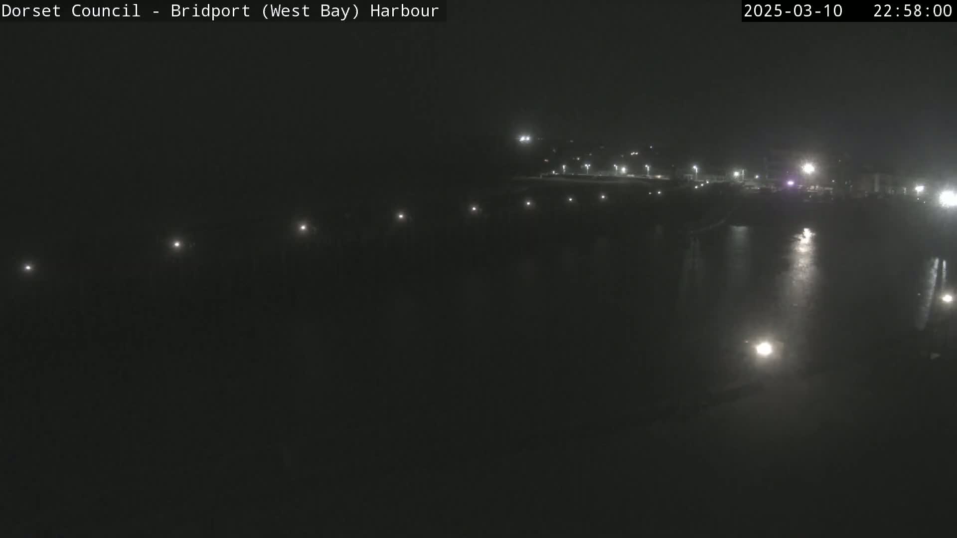A nighttime harbor scene with calm water reflecting lights from the shore.