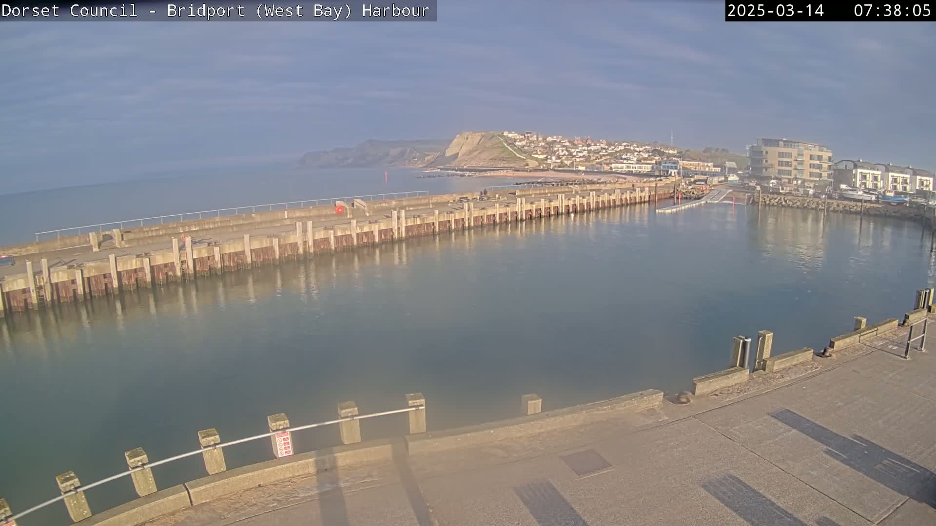 A harbor with a long concrete pier extends into calm, clear water under a mostly sunny sky, with a town and cliffs visible in the background.