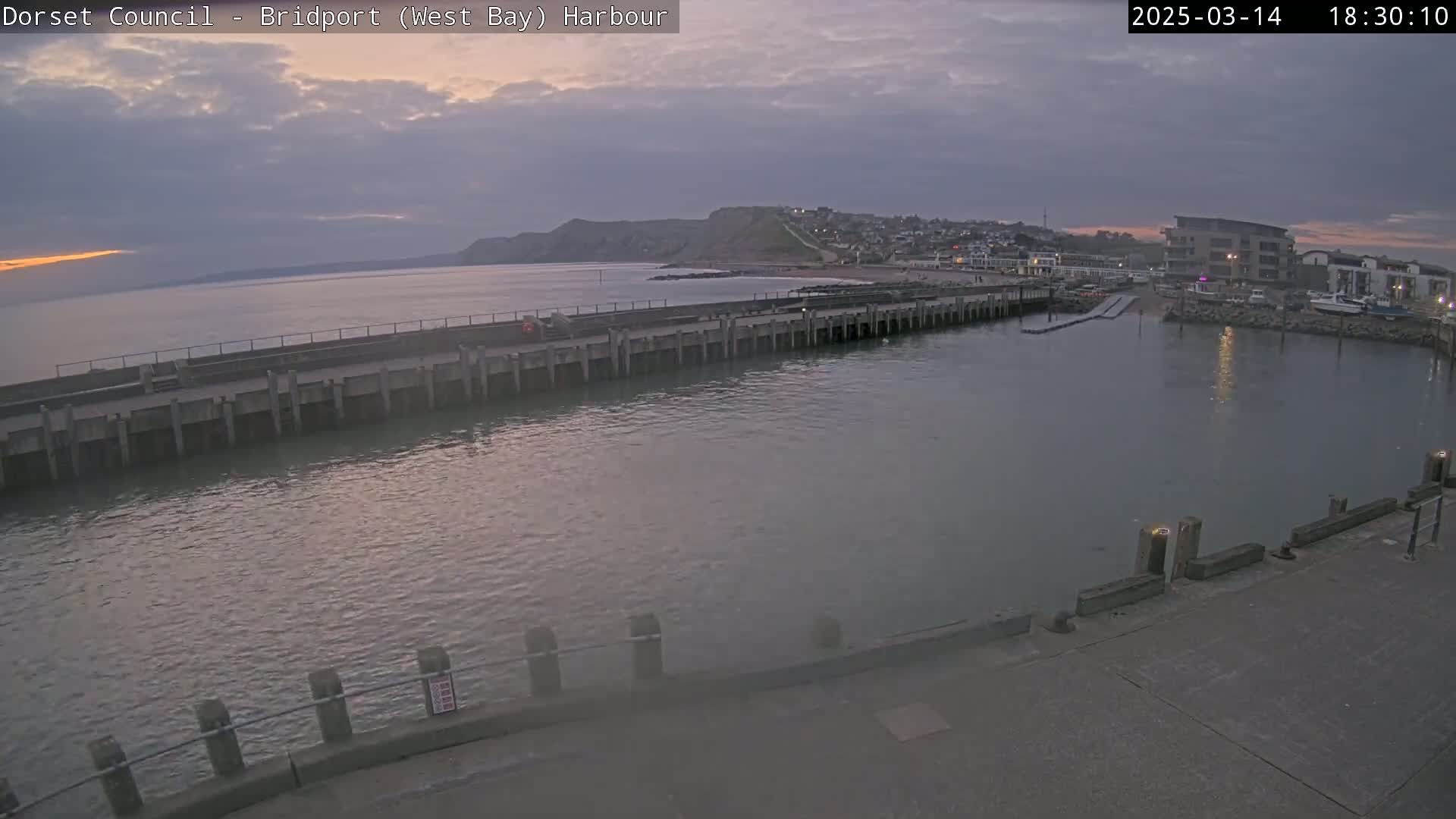 A harbor at dusk shows a long pier extending into calm water, with a town and a hill visible in the distance under a cloudy sky.