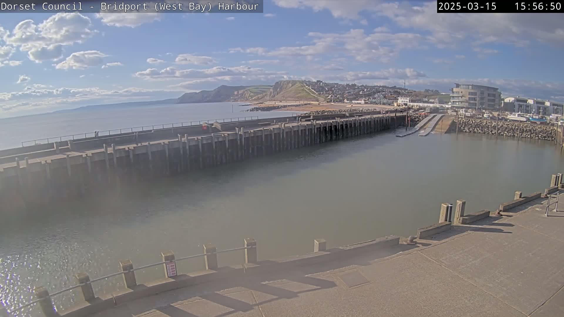 A harbor with calm, light-brown water is seen under a partly cloudy, sunny sky, with a long concrete pier extending into the water, and a town and cliffs visible in the distance.