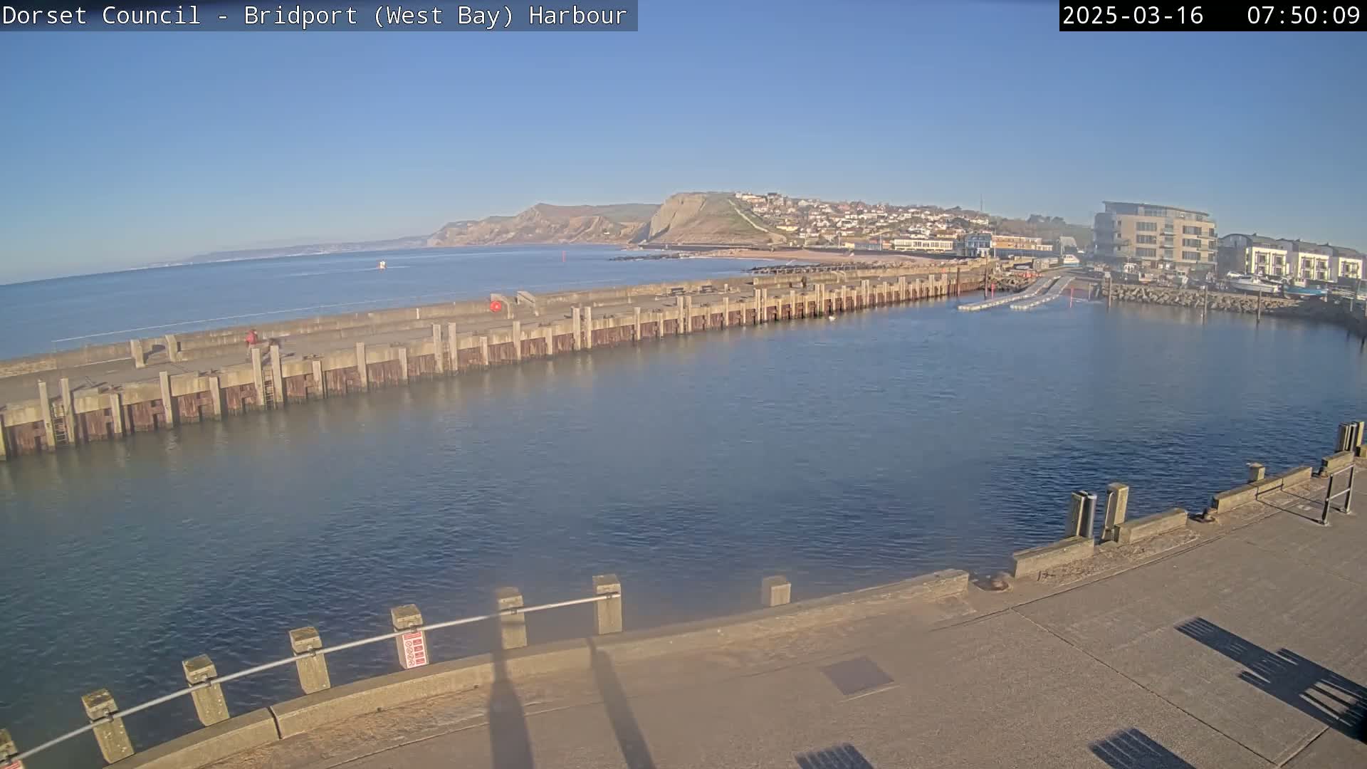 A harbor with a long concrete wall separating the calm water from a beach and a town under a clear, sunny sky.