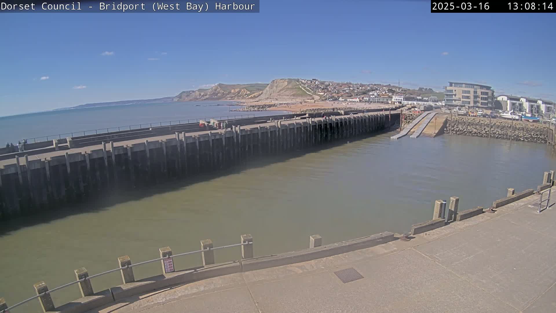 A harbor with calm water and a long concrete wall separating it from a sandy beach under a sunny sky.