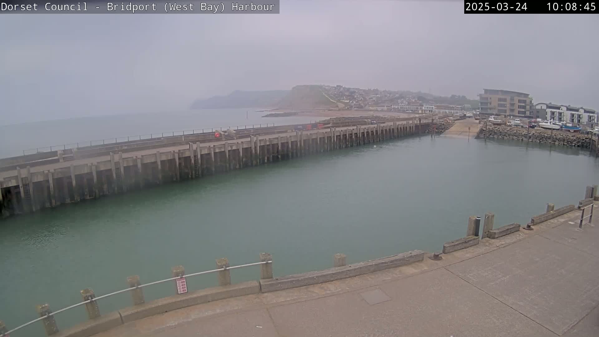 A harbor with a long concrete pier and calm, greenish water is seen on an overcast day, with a small town and a hillside visible in the distance.
