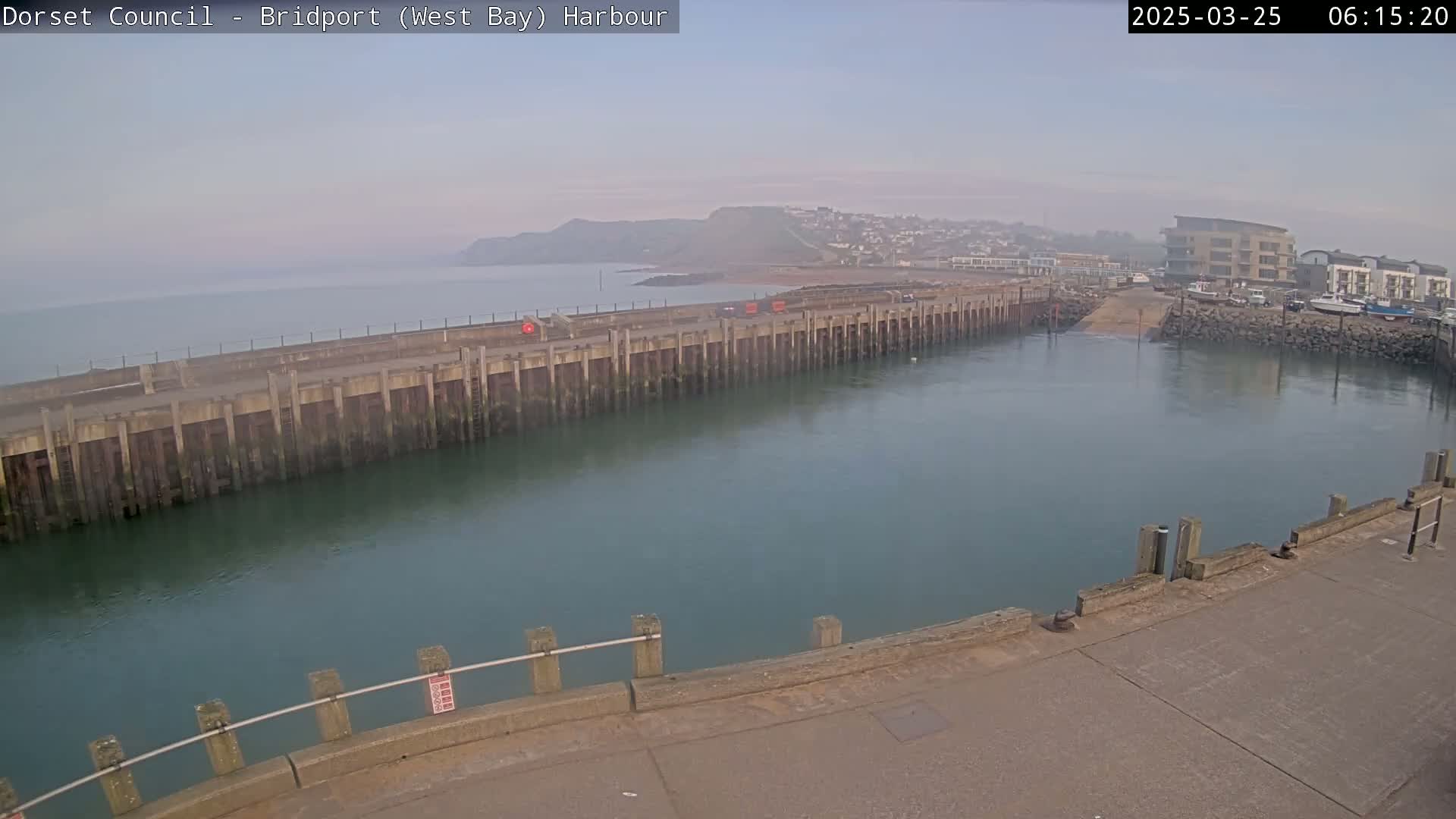 A harbor with a long concrete pier juts into calm, greenish-blue water under a hazy sky.