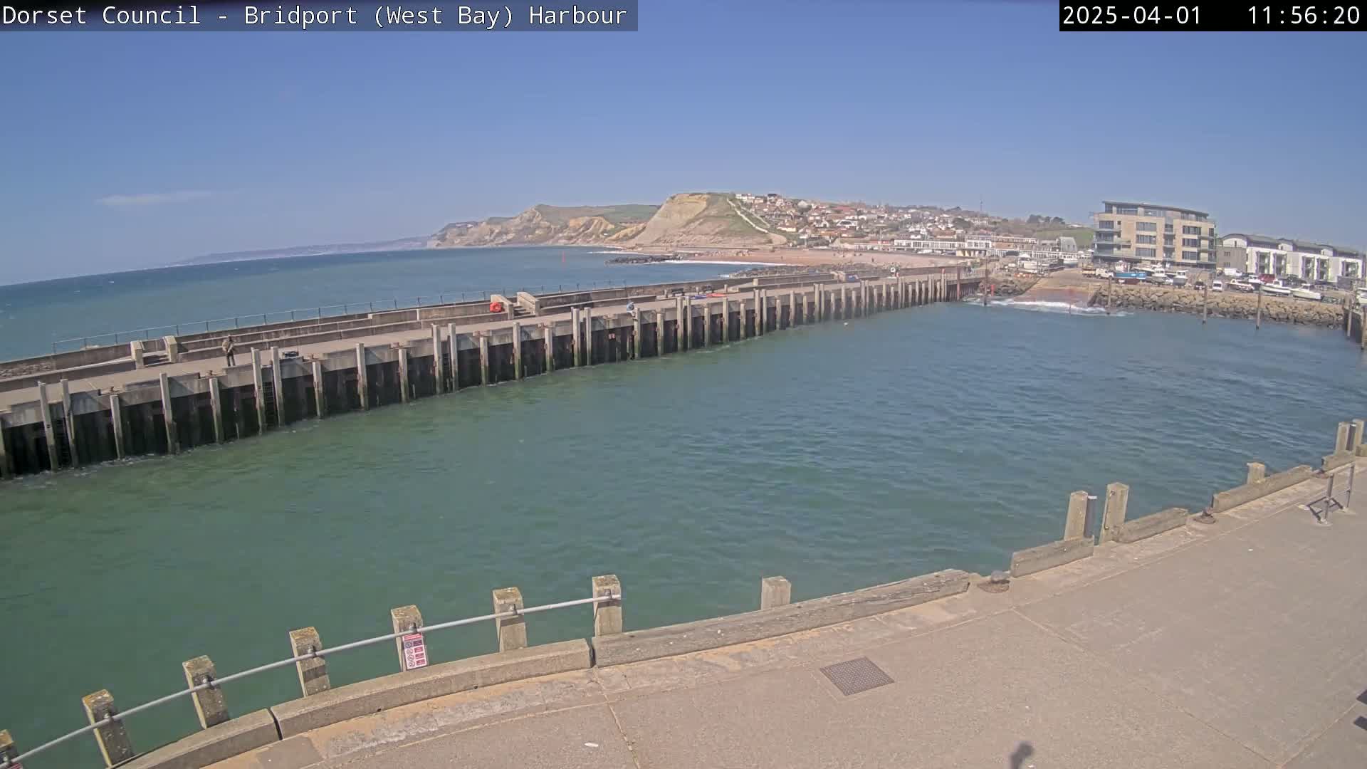 A harbor with a long pier extending into calm, green water under a clear blue sky, and a town and cliffs visible in the distance.