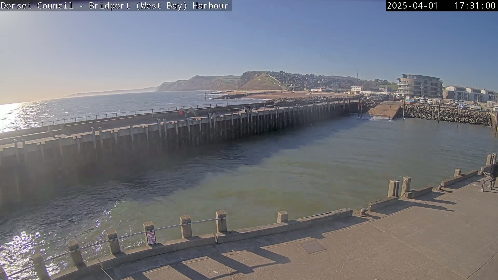 A harbor with a calm sea under a sunny sky, featuring a long concrete pier extending into the water, a small boat ramp, and a town visible in the distance.