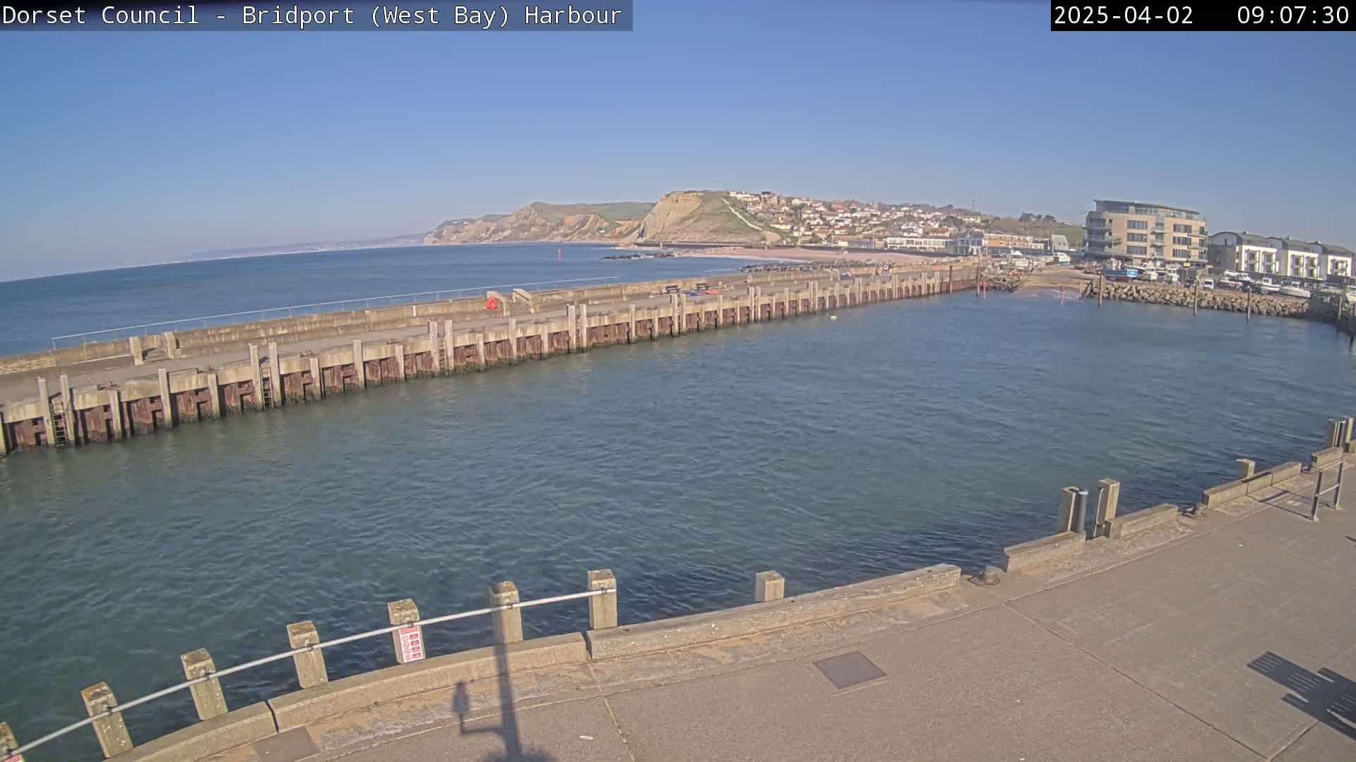 A harbor with a long concrete pier extends into calm, blue water under a clear blue sky, with a town and cliffs visible in the distance.