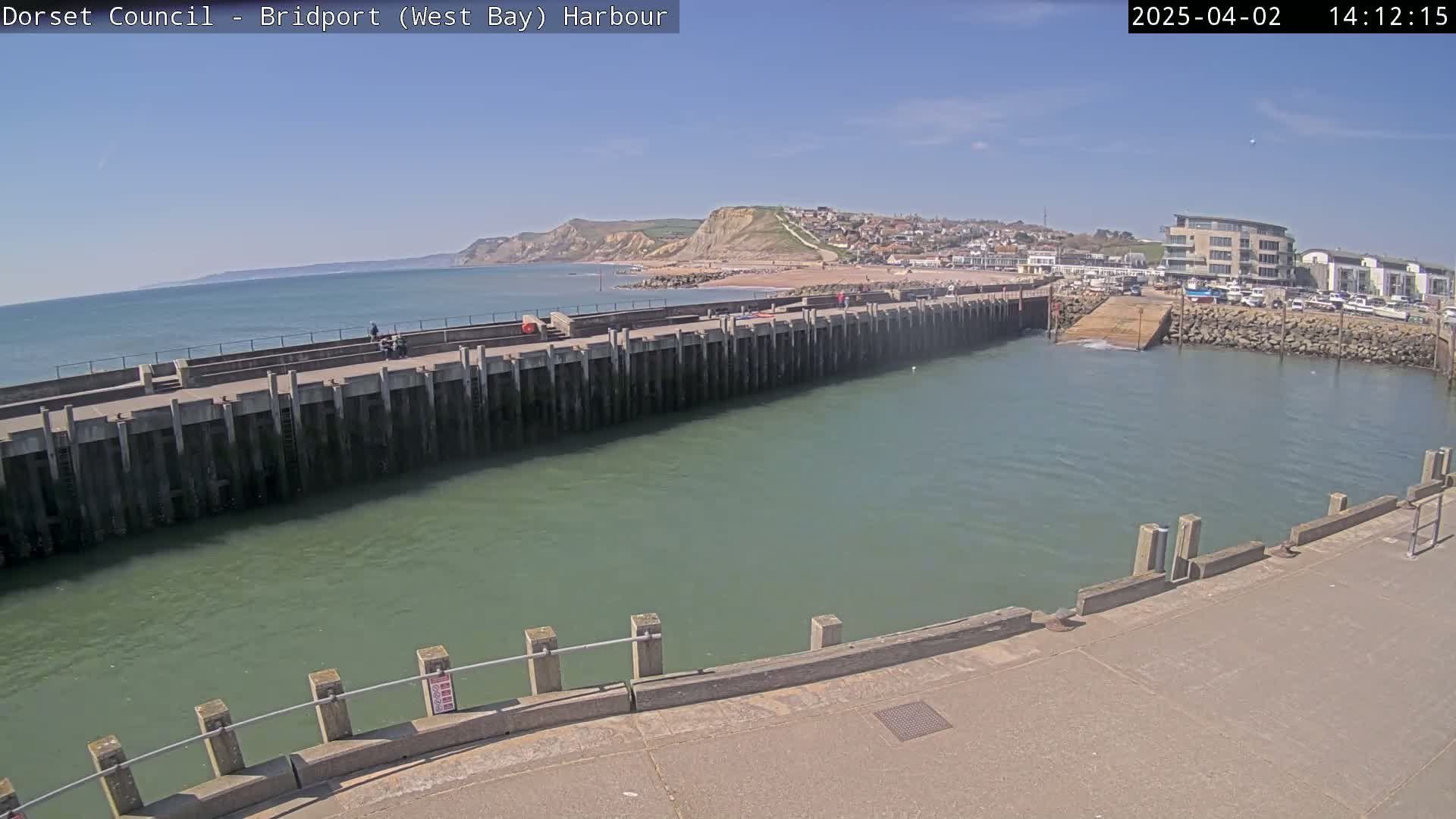 A harbor with a long concrete pier extends into a calm, greenish-blue body of water under a clear, sunny sky, with a sandy beach and cliffs visible in the distance.