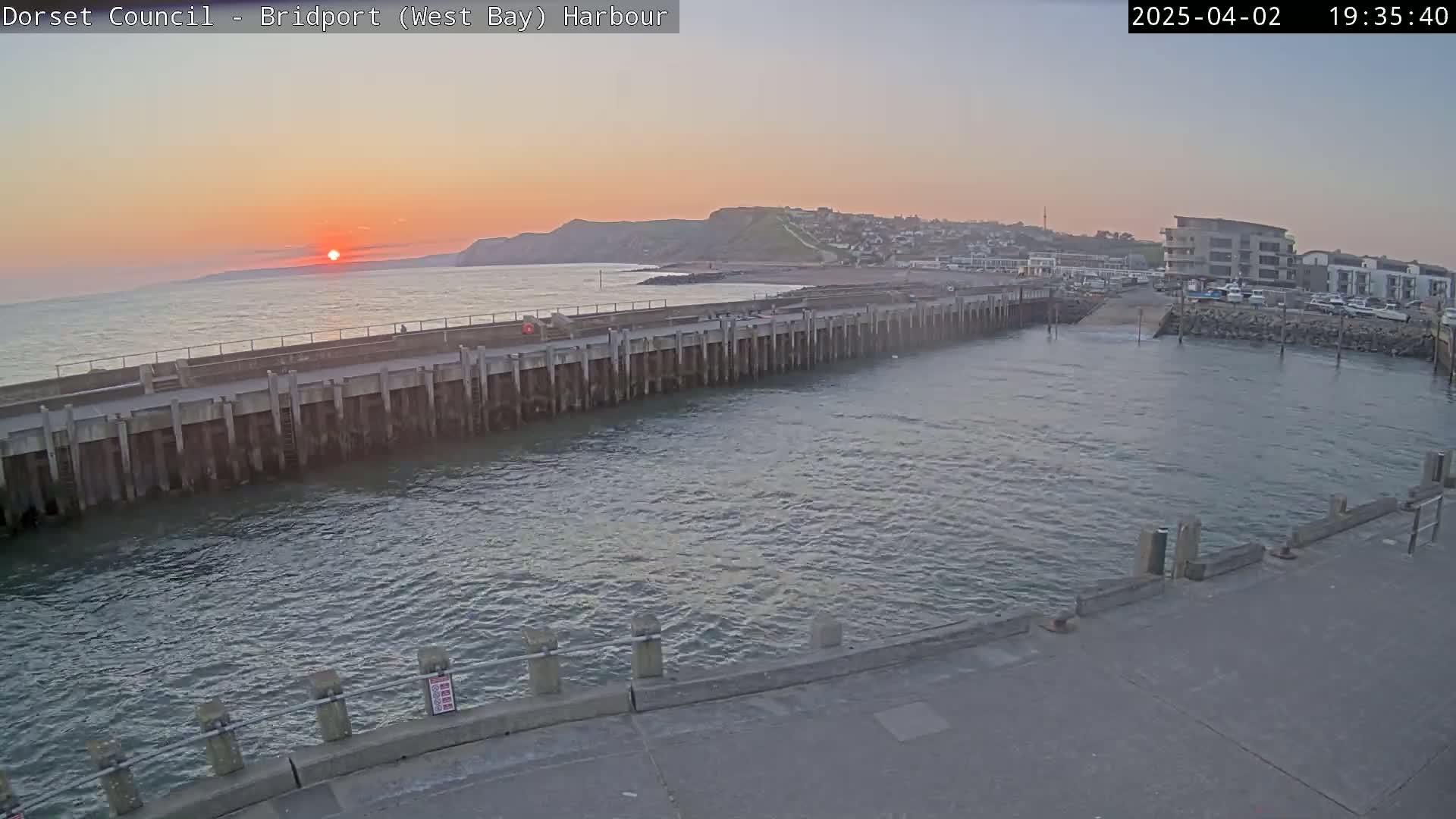 A harbor at sunset, with calm water reflecting the orange and pink sky, shows a long concrete wall separating the harbor from the open sea and a small town nestled on a hillside in the distance.
