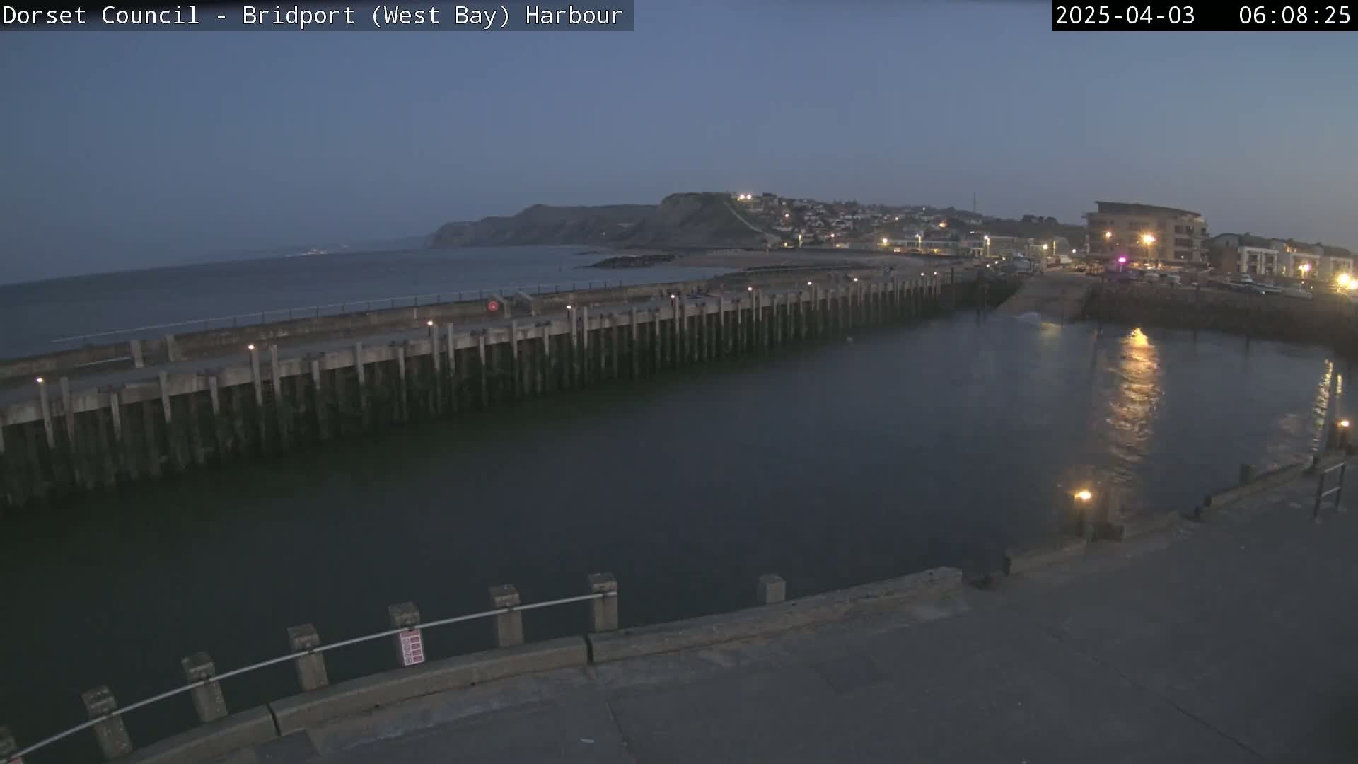 A harbor at twilight shows a calm body of water next to a concrete pier, with a distant coastline and a town visible under a dark, clear sky.