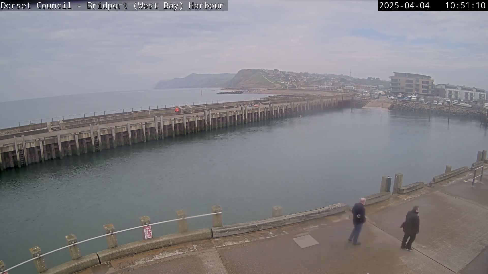 A harbor with a long concrete pier extending into calm, overcast waters, two people stand on the pier, and a distant coastline is visible.